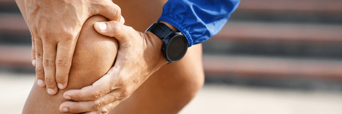 Person wearing a blue sleeve holding and supporting their knee, indicating knee pain or injury during activity in a close-up shot of hands on the joint.